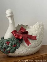 Front view of white ceramic swan plant holder with red bow and green holly leaves with red berries, detailed sculpted feathers and scalloped edge opening.