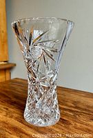 Single photo showing a clear crystal vase with a flared top and detailed pinwheel pattern cut into the glass. The vase sits on a wooden surface with a neutral background.