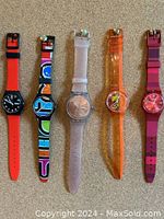 Five Swatch watches laid flat on surface showing colors and strap styles