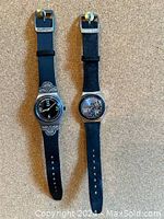 Two metal wristwatches displayed side by side on a carpeted floor background. The left watch has a cloth strap and decorative silver design around the face. The right watch has a leather strap and floral design on the dial.