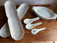 Top view of 3 long white ceramic serving trays with scalloped edges and 4 white ceramic soup spoons on a wooden surface.