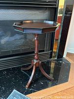Full view of the wooden wine serving table with octagonal tray top and pedestal base in front of a black fireplace.
