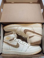Side profile of pair of Air Jordan 1 Retro High OG sneakers in cream leather with cork-textured accents, sighted inside original box.