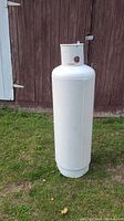 Full view of the tall white propane tank standing upright on grass in front of wooden wall.