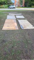 Five plywood sheets arranged outdoors on grass surface, showing different sizes and conditions including nailed wood and metal strip