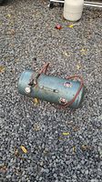 Vintage cylindrical air compressor tank laying on gravel showing pressure gauge and attached orange hose