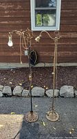 Two full-length antique metal floor lamps standing outdoors on pavement, with visible cords and bulbs. One has a decorative scroll arm with bulb socket, the other has a curved arm with a lampshade holder.