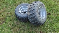 Two ATV tires mounted on metal rims resting on grass. Tires show aggressive tread and wear.