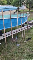 Photo showing six garden tools leaning against a boat, including pitchforks, hoes, and rakes having wooden handles and metal working ends.