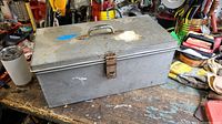 Closed grey metal tool box showing latch and handle on lid, placed on a workbench with various tools in the background.