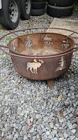 Side view of round metal fire pit with moose, tree, and deer cutout designs. Shows rust and weathering.