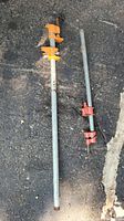 Two pipe clamps laid on asphalt, one with orange hardware and one with red hardware.