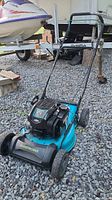 Full side view of Yardworks push mower on gravel with boat trailer in background, showing mower deck, handle, and wheels.