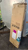 Large cardboard box containing the MAAX shower door, standing upright outdoors next to a trailer.