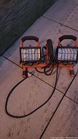 Two halogen work lights with protective metal grills in orange frames, shown plugged in and off, laying on a tiled floor.