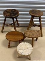 Photo showing lot of five wooden stools of various shapes and finishes on a neutral floor with a metallic background.