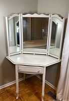 White corner vanity table with three-panel mirror in a room corner, wooden parquet floor, showing front and angled views.
