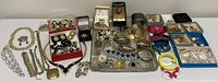 Full layout of assorted costume jewelry displayed on a white surface including necklaces, bracelets, brooches, earrings, and hair clips spread over trays, boxes, and loose.