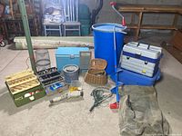 Full lot image showing the Coleman cooler, 55-gallon barrel, blue water catcher/food storage, two tackle boxes, fishing creel basket, nets, ice auger, rope, and hatchet.