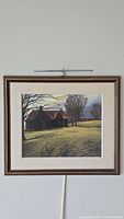 Framed pastel artwork showing entire country house scene with trees and grassy field, wall-mounted with picture light above.