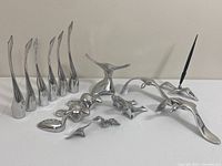 Full group view of 6 tall Hosleton crane sculptures, 5 smaller bird figurines, and 2 aluminum bird pen holders with black pens placed on white surface against beige wall.