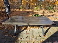 Full view of rectangular outdoor table on stone patio with autumn leaves around. Table has slatted top and central umbrella hole with metal legs.