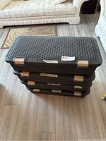 Stack of 4 black plastic rolling storage bins with woven pattern, each labeled with contents such as towels and flannel sheets, on a wooden floor in a living room setting.