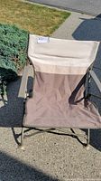 Front view of folding camping chair showing beige and brown fabric with metal frame and armrests, situated outdoors on a concrete surface under sunlight.