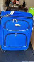 Front view of blue Delsey spinner carry-on suitcase showing front zippered compartments, Delsey logo badge, and spinner wheels.