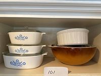 Three CorningWare casseroles with lids stacked on the left, all with blue floral Cornflower pattern. On the right, three nested Pyrex bowls in graduated sizes with a brown to amber gradient exterior and a white ceramic ribbed round baking dish stacked on top.