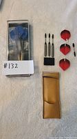 Photo of one dart set with three metal darts and blue flights inside a clear plastic case, with extra red flights and a gold pouch next to it.