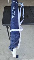 Full view of the Nautica table umbrella in its navy blue protective cover with the Nautica logo printed in white vertically along the length of the cover. Attached is a white strap for carrying.