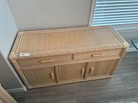Front angle view of beige rattan style credenza with woven rattan drawers and cabinets, sitting against wall near window on wooden floor.