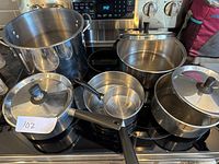 Image showing six stainless steel pots on a stovetop, some with lids and black handles, varying in size.