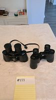Two black pairs of binoculars on a beige surface with attached neck straps and lens caps partially present.