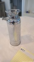 Full view of the silver-tone metal cocktail shaker shaped like a small fire extinguisher, standing upright on a light surface.
