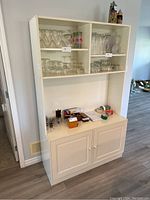 Front view of large white wooden cabinet showing open upper shelves and lower double doors with knob handles. Some small items are placed on the top surface but contents not included in sale.