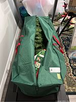 Photo of a green zippered carrying bag partially open to reveal an artificial Christmas tree inside, along with a tree skirt with Christmas designs.