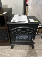 Front and slightly angled view of a black Komodo electric fireplace model SP5295 with door closed and manual placed on top.