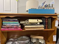 Wooden shelf with books including cookbooks and piano books, blue file organizer on top shelf, assorted kitchenware on bottom shelf