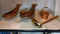 Photo showing two large boat-shaped copper dishes, small embossed copper item, kettle, bowl, and ladle on display with white cloth background.