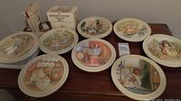 Eight decorative plates arranged on a table featuring Beatrix Potter characters, small books stacked at the back, and decorative bookends visible.