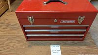 Closed red metal Craftsman tool box with black handle and silver latches on wood surface.