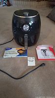 Black Dash electric air fryer on floor with air fryer liners box and instruction manual next to it.