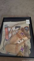 Shadow box containing a pair of tan leather gloves with fur trim and floral beadwork, along with a painted hide with Indigenous designs.