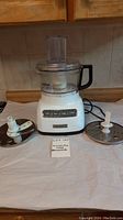 Full view of KitchenAid food processor base unit with clear bowl and lid, four main components shown including blades and the note 'KitchenAid food processor'