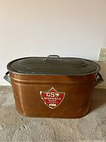 Photo of the copper boiler with lid closed, showing the GSW Monarch Ware logo on front and two black handles on sides.