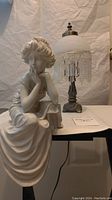 Greek style lady figurine seated with detailed flowing dress and head resting on hand alongside small beaded table lamp