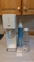 White SodaStream carbonation machine, blue CO2 cylinder, and transparent plastic bottle with white cap and base displayed side-by-side on kitchen countertop.
