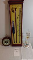 Photo showing full storm glass with thermometer in wooden frame, German barometer, and Galileo thermometer on a table.
